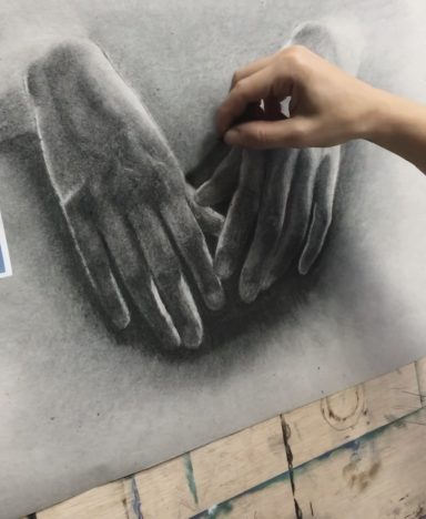Manos grabándose en un dibujo a lápiz, una sobre la otra, en un fondo difuminado.